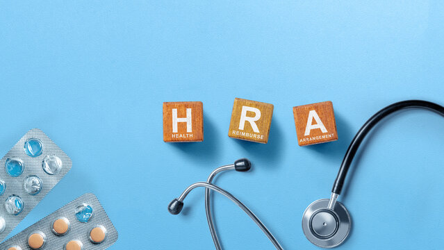 Wooden blocks spelling HRA for Health Reimbursement Arrangement alongside a stethoscope and pills on a blue background. Concept of Reimburse