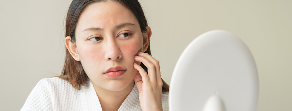 Asian young woman looking face skin in the mirror have a red rash on her face from cosmetic allergy