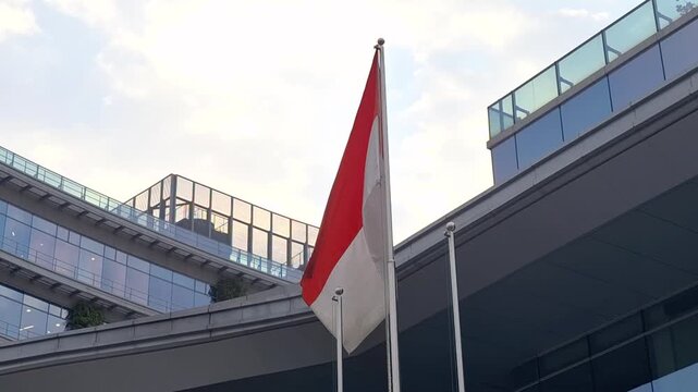 Red and white flag waves in front of modern glass building. Sky shows soft clouds above the structure. Flagpole stands tall beside sleek architectural lines