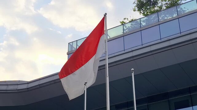 Red and white flag waves in front of modern glass building. Sky shows soft clouds above the structure. Flagpole stands tall beside sleek architectural lines