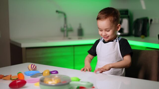 Smiling boy in apron playing with toy food at table. A happy child sits in a modern kitchen with green lighting while interacting with colorful plastic dishes.
