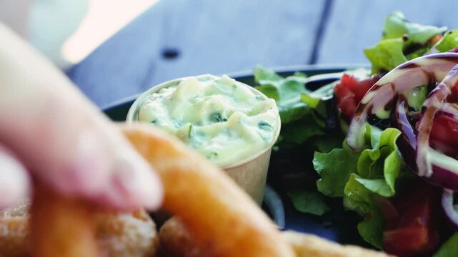 Close up of dipping a crispy onion ring into creamy sauce with fresh salad on the side. Appetizer snack and restaurant dining concept. High quality 4k footage