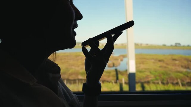Silhouette of a passenger recording a voice message on a smartphone while sitting by the train window. Mobile communication during travel and modern lifestyle concept. High quality 4k footage