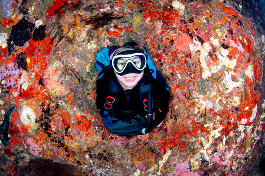 Asian female scuba diver with a smiley face posing through the natural circle hole underwater. Colorful hard coral cover on the rock around the scene.