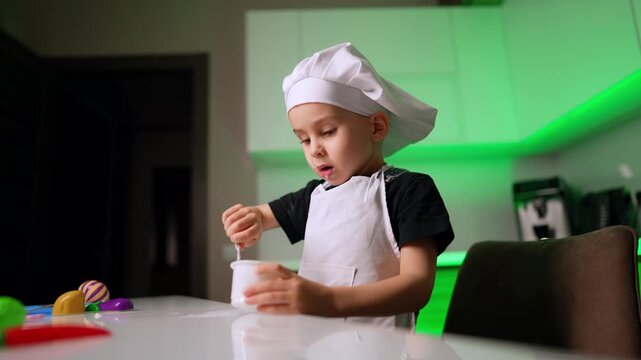 Child chef stirring food in plastic cup at table. A focused young boy wearing a chef hat uses a spoon inside a small container in a green illuminated kitchen.