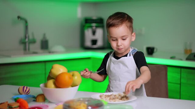 Little boy in apron holding fork over plate of food. A young child eats a meal at a white table next to a fruit bowl filled with apples and an orange in a green kitchen.