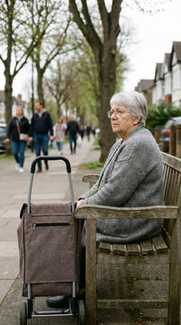Solitary elderly woman seated on a street bench beside a worn shopping trolley