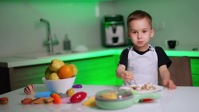 Young boy in apron eating food with fork at table. A child sits at a white table next to a bowl of fresh fruit while holding a fork over a plate of food in a kitchen.