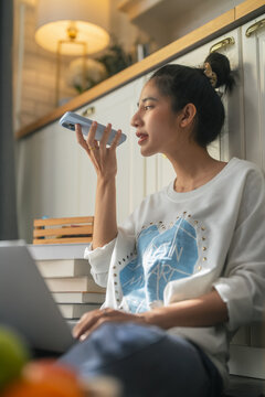 A woman speaks into her smartphone using a voice command feature, sitting comfortably in a warm, softly lit kitchen nook surrounded by books and cozy home decor, blending everyday life with smart tech