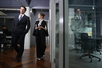 Fototapeta premium Tall bearded manager laughs at his colleague comment while they walk, genuine amusement breaking across his face in the after hours office corridor in a bright modern coworking office.