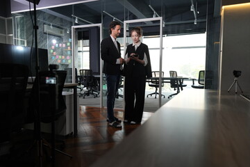 Behind the scenes, a bun-haired woman shows a handheld device to a crew member amid tripods in a glass-partitioned coworking office at dusk, talking softly in the ambient evening light. © ultramansk