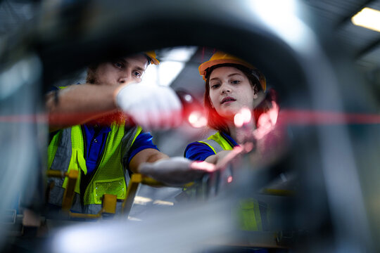 Bearded male warehouse worker and young female colleague in hard hats and safety vests point a barcode reader at components between machine frames at an automotive parts production plant.