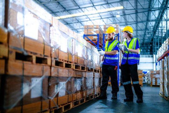 Two warehouse employees, a bearded man and a young woman, wearing yellow hard hats point out carton labels that need rechecking in a spacious logistics centre filled with shrink wrapped.