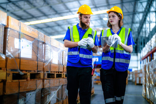 Bearded male warehouse worker and young female colleague in hard hats and safety vests scan barcode labels on a shrink wrapped pallet in a spacious logistics centre filled with shrink.