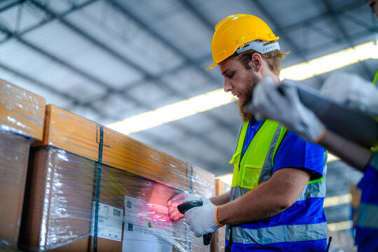 Male warehouse supervisor with a beard and his female coworker in protective workwear aim a glowing barcode reader at stacked cartons in a bright modern distribution warehouse with tall pal.