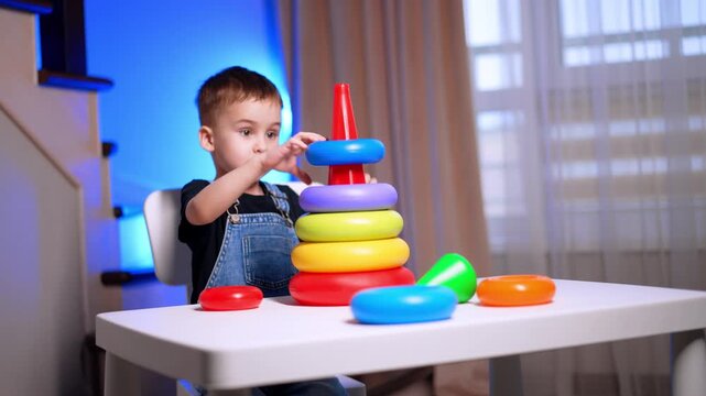 Boy stacking colorful rings on cone toy. He reaches toward a blue piece while seated beside the white play table.