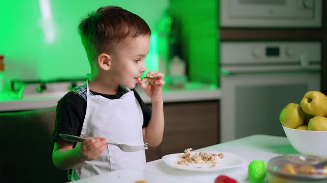 Side view of boy in apron eating with hand. A young child holds a fork while using his fingers to eat food from a white plate in a green illuminated kitchen.