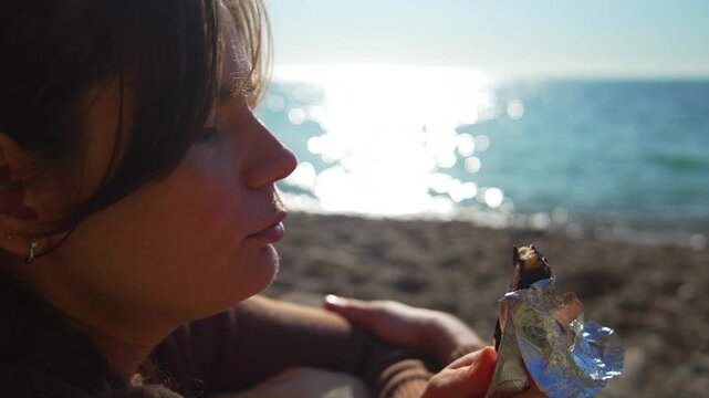 Woman eating chocolate as she sits on a sunny pebble beach with sparkling sea water in the background