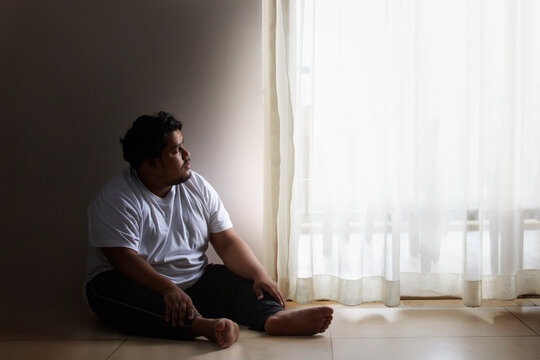 A fat man sitting sadly looking at the curtains in his room
