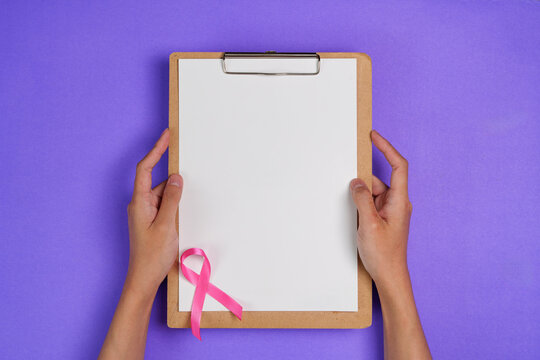 Breast cancer awareness concept with pink ribbon on blank clipboard held by hands over purple background