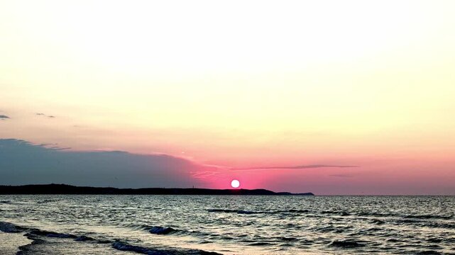Wide tranquil ocean view at sunset with calm horizon, distant coastline, gentle waves, soft reflections and warm gradient sky shifting through pink, orange, yellow and blue tones, with sunset