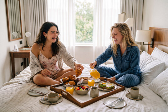 Two young women share a joyful hotel room breakfast in bed with croissants, fruit, orange juice, and avocado toast.