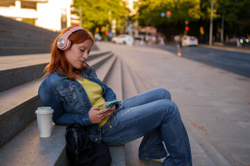 Fototapeta premium Young woman relaxing on city steps, listening to music with pink headphones and using smartphone