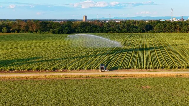 Irrigation sprinkler watering field near wind turbines. Parallel crop rows extend toward trees and distant industry beneath scattered clouds.