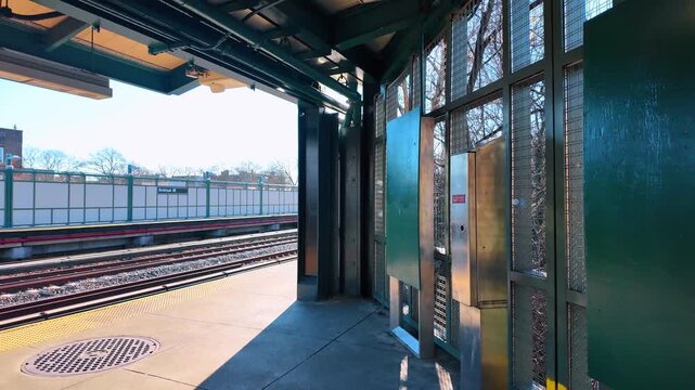Subway station platform at Avenue M in New York. Bright sunlight casts long shadows across an outdoor train platform bordered by green mesh panels.