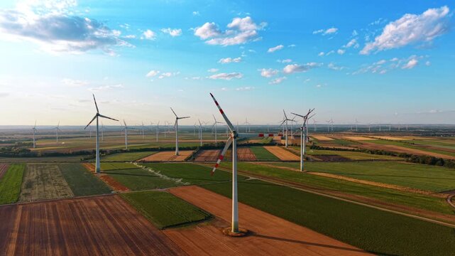 Aerial view of wind farm across agricultural fields. Rows of turbines span green and brown farmland beneath a bright clouded sky.