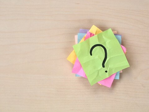 Handwritten question mark on a stack of colorful adhesive notes on wooden desk background with copy space. Concept for questions, FAQ, problem solving, confusion, uncertainty, and decision 