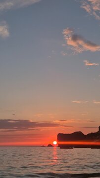Ocean sunset, golden sun dips behind a coastal cliff at dusk while gentle waves roll ashore. Static wide shot.