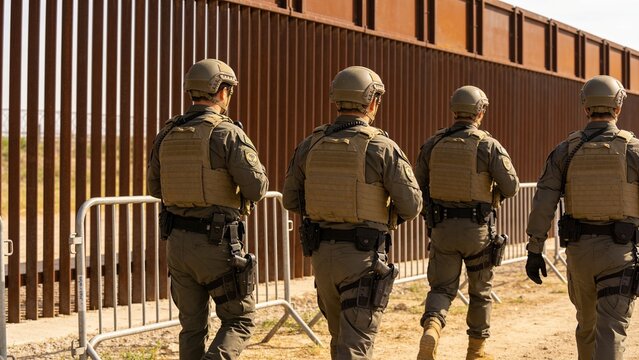Armed border patrol agents walking along high security metal fence in sunny desert area.