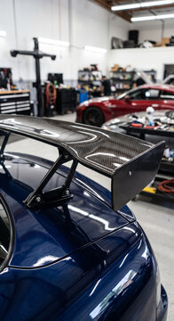Carbon fiber rear wing and glossy trunk section on a blue modified sports car in a garage. Clear lighting emphasizes its fitment, surfaces, and functional performance design.