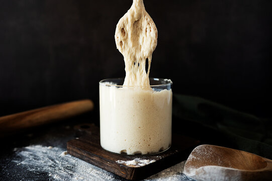 Wooden spoon lifting active gooey sourdough starter from a jar.