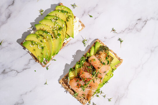Two healthy crispbread sandwiches with avocado and salmon under bright sunlight.