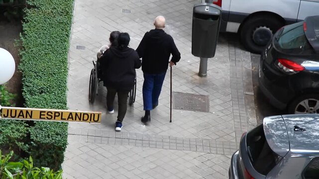 Viejo con bast&oacute;n, silla de ruedas y cuidadora residencia empujando en ayuda en paseo y andar movilidad y asistente social por calle de adoqu&iacute;n, arbusto y coches aparcados