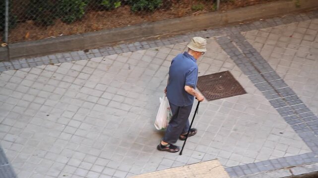 Abuelo con bast&oacute;n, bolsa de compra y sombrero paseando lento en vida activa saludable paseo con sandalias cargado por acera calle tercera edad viejo andando en adoqu&iacute;n urbano