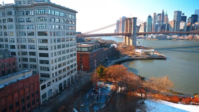 DUMBO waterfront beside Brooklyn Bridge. Historic buildings overlook the East River and a snow covered park in winter sunlight.