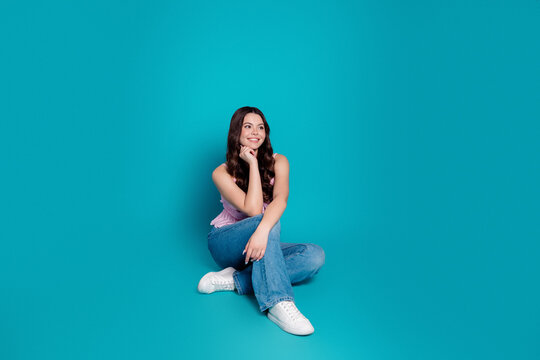 Young woman sitting on studio floor smiling and looking away against turquoise background in casual jeans and white sneakers, relaxed thoughtful portrait conveying optimism