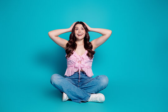 Young woman sitting cross-legged on turquoise background looking surprised and happy in a pink top and jeans