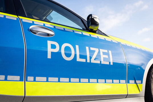 Close-up of a German police car door featuring the "Polizei" lettering in white on the iconic blue and neon yellow design against a bright sky.