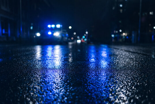 Blue emergency lights from a police vehicle reflect dramatically on a wet urban street at night, creating an atmospheric and moody scene.