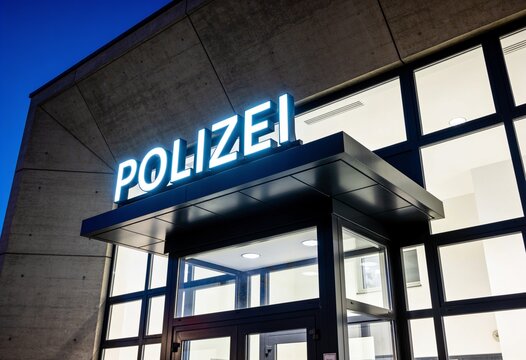The entrance of a German police station at night with a glowing "Polizei" sign above the modern glass door canopy.