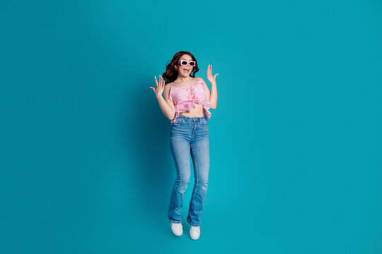Joyful young woman jumping in casual outfit with pink top and sunglasses against a bright blue background