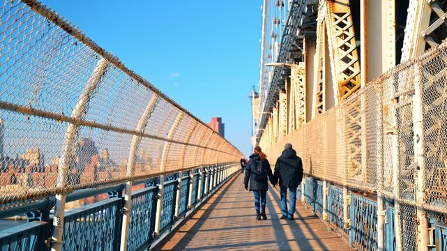 Pedestrians walking across Manhattan Bridge walkway. Two bundled figures recede between chain link fences in warm low sunlight.