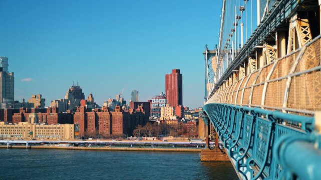 Manhattan Bridge railing overlooking Lower Manhattan. Sunlit steelwork leads toward dense waterfront buildings under a cloudless blue sky.