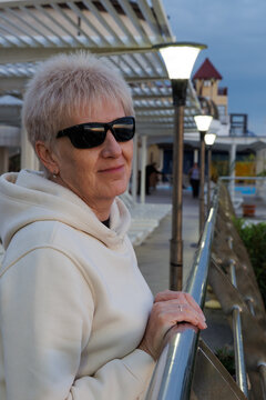 Smiling mature female in sunglasses and casual hoodie leaning on metal railings at modern coastal resort terrace in evening.
