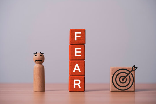 Close-up of wooden blocks spelling FEAR beside a worried figure and a target icon, representing overcoming fear, achieving goals, building confidence, personal growth and success.