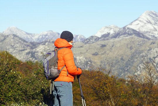 Hiker faces snow-capped peaks through autumn shrubs on Vrmac, Bay of Kotor. Conveys first-snow seasonal contrast, autumn-to-winter mountain transition, and solitary hiking above the Adriatic coast.